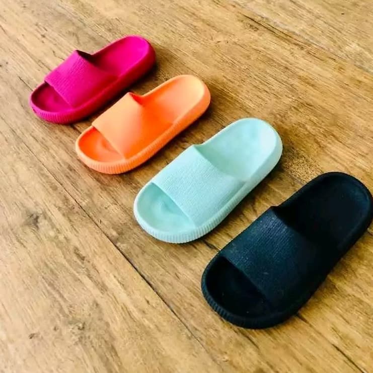 Quatro chinelos 'nuvem' infantis em cores rosa, laranja, turquesa e preto sobre uma superfície de madeira.