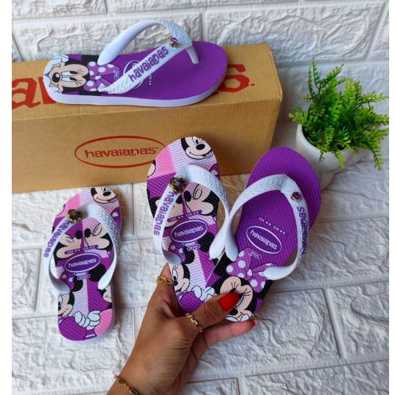 Sandálias infantis com estampa de Minnie Mouse, cores roxo e rosa, seguradas por uma mão.