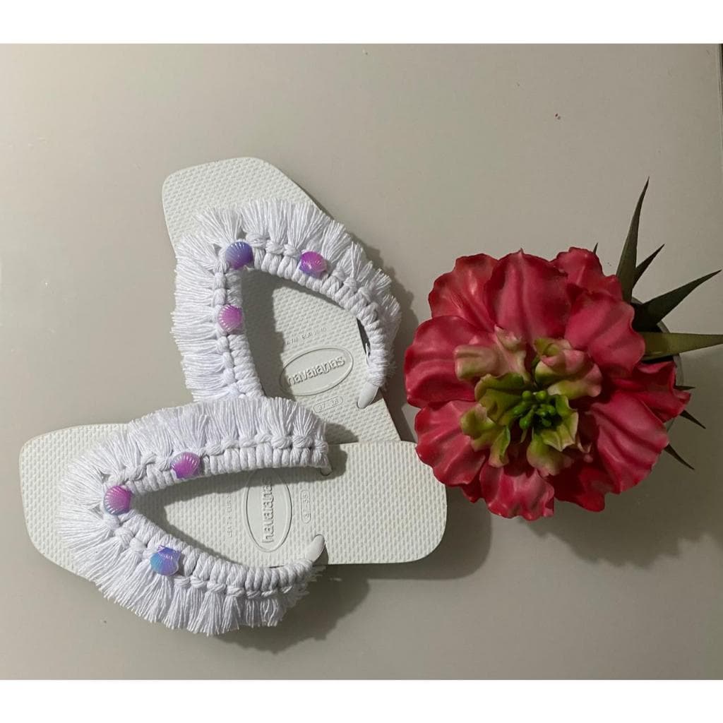 Chinelos Havaianas customizados brancos com pedrarias e franjas, ao lado de uma flor rosa