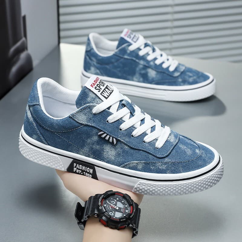 Tênis masculino azul com estampa de jeans, estilo casual, segurado por uma mão, mostrando detalhes da sola e cadarço brancos.