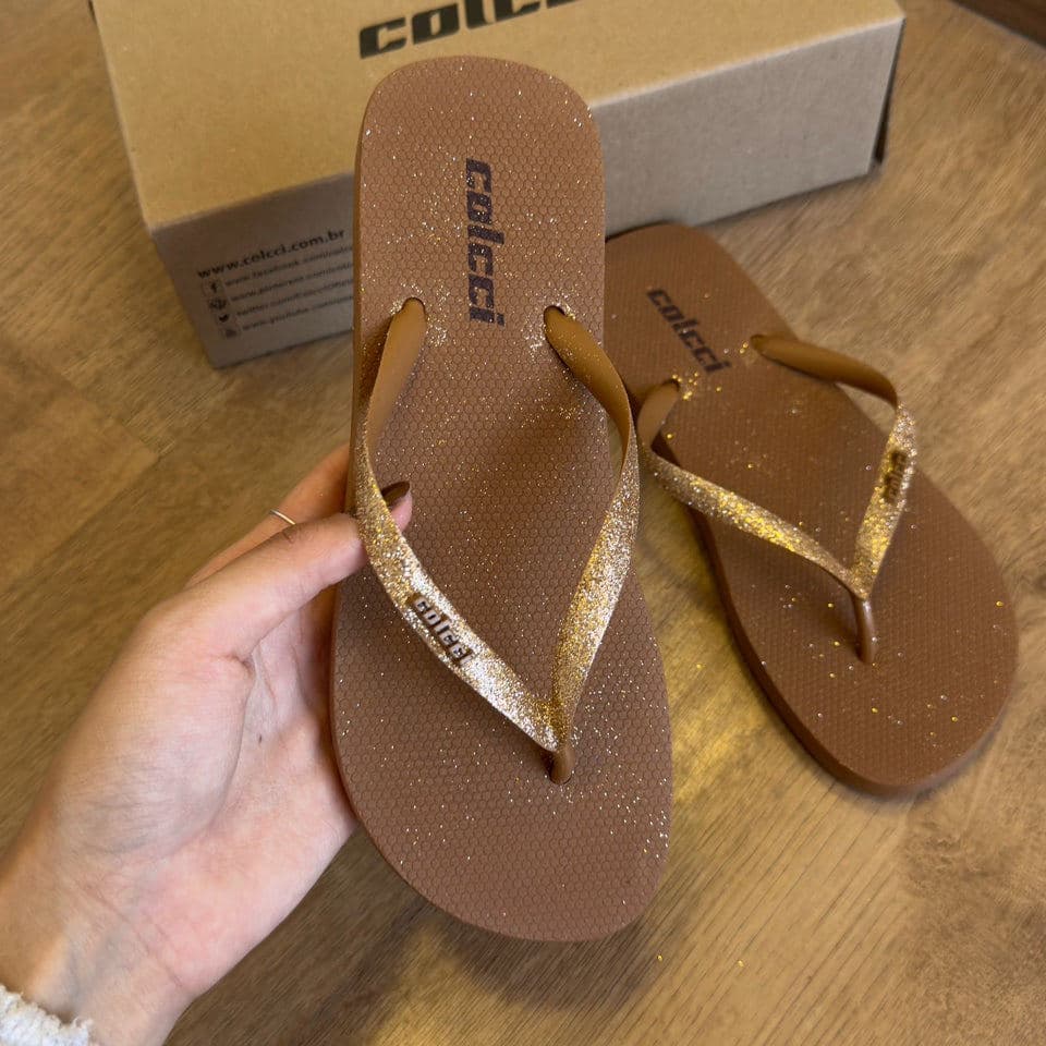 Chinelos de dedo Colcci marrom com glitter dourado, segurados por uma mão, com uma caixa de fundo de madeira.