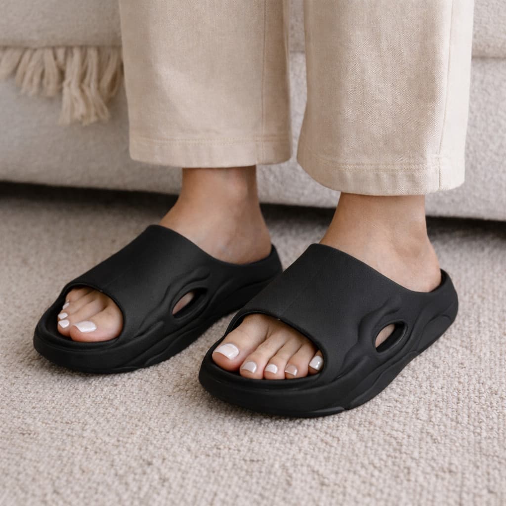 Chinelo slide preto anatômico com ventilação, usado por uma pessoa com unhas pintadas de branco.