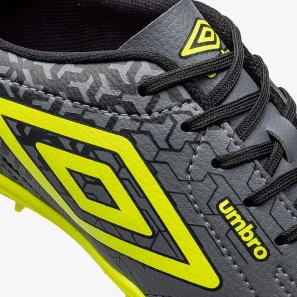 Detalhe do tênis Umbro Class Neo para futebol, mostrando o logotipo e design em preto, cinza e amarelo.